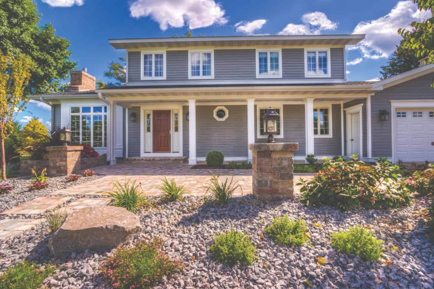 A modest two-story home with gray siding and matching white trim around every window. The front yard is paved with paver stones, gravel, and plants and the house stands against a bright blue cloudy sky.
