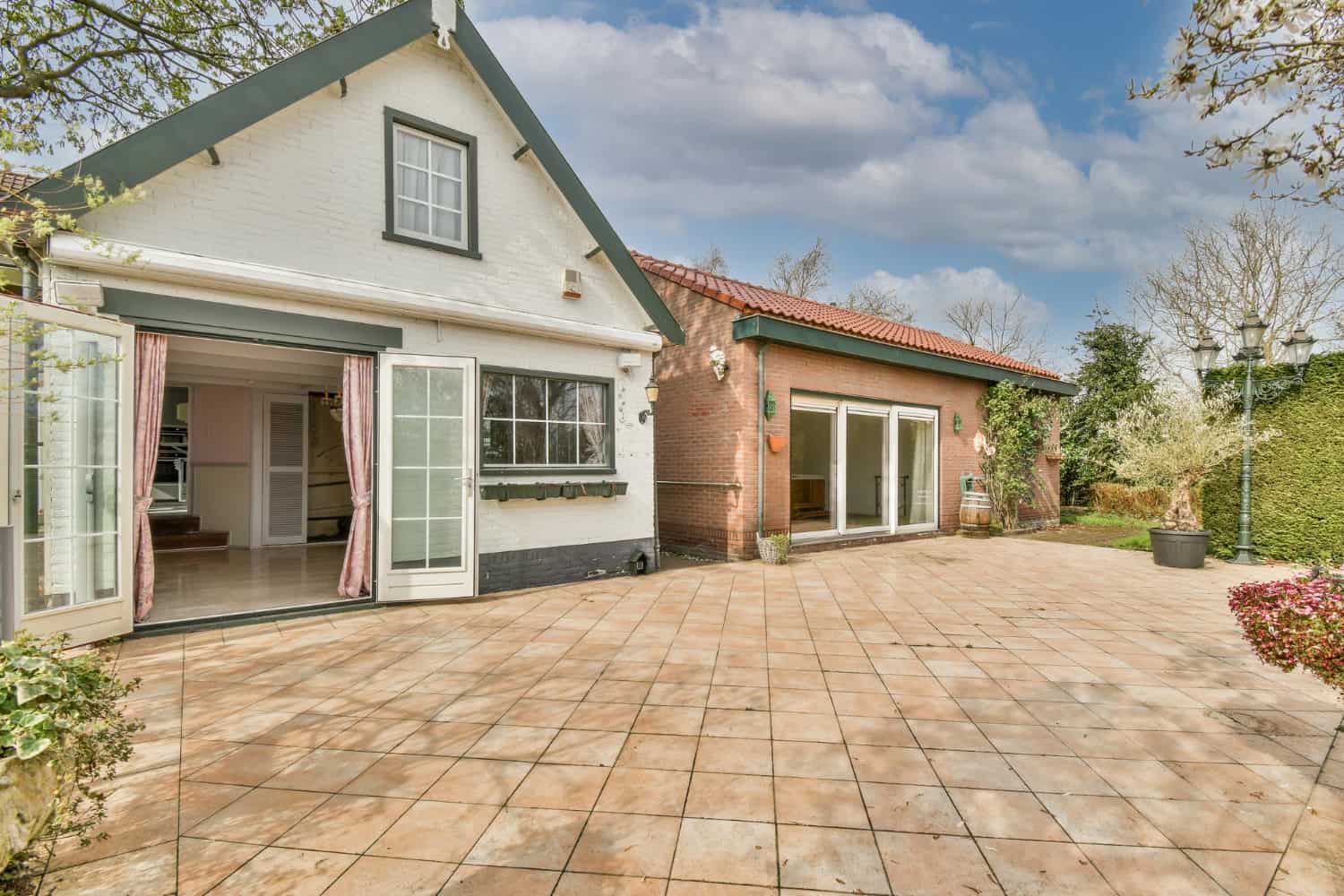 The back patios of two side by side white and tan brick homes; a white brick home has double French patio doors propped open. The second tan brick house has three panel sliding patio doors.