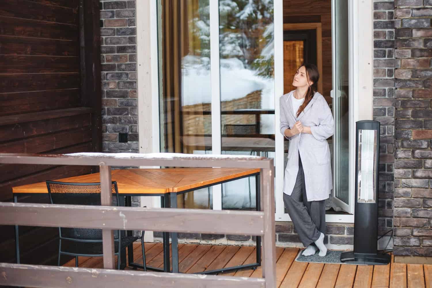 A woman wearing a light gray house robe and dark gray sweatpants stands just outside her patio door on a wooden deck on a cold winder morning. There's a wooden table and chairs, and a snow covered railing.