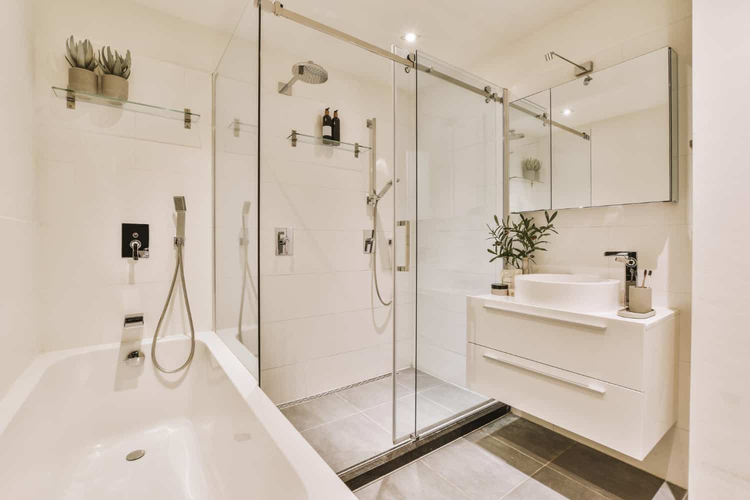 AN elegant all-white bathroom with a large glass door shower and whiter tub. The walls are covered with white tiles and recessed lighting makes the room bright and well-lit.