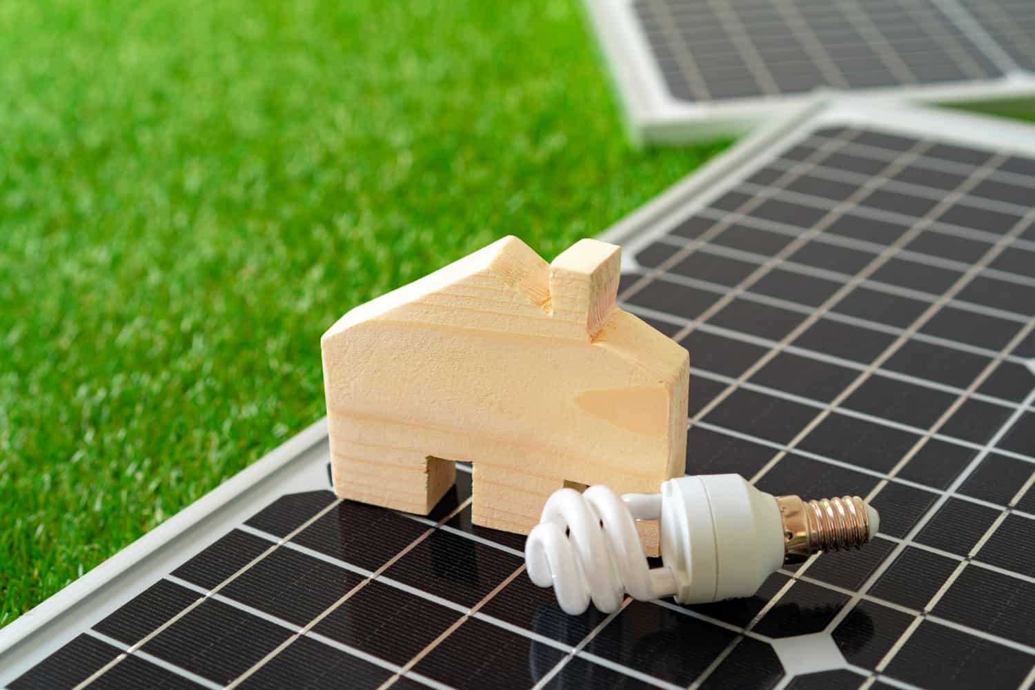 A small piece of wood in the shape of a house sits on top of a solar panel and next to an energy-efficient LED lightbulb. There is green grass in the background.